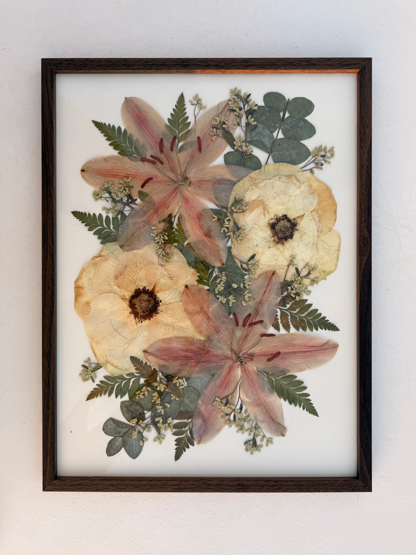 Pressed pink lilies, ivory roses, fern and eucalyptus on ivory paper in a dark chestnut frame